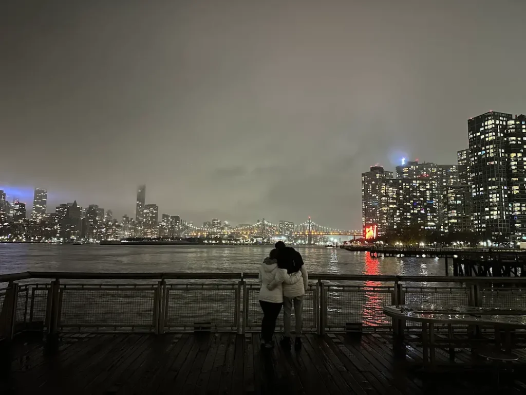Photo d'une mère et son ado, bras dessus bras dessous, devant la skyline illuminée de nuit, à Gantry Plaza State Park, New York