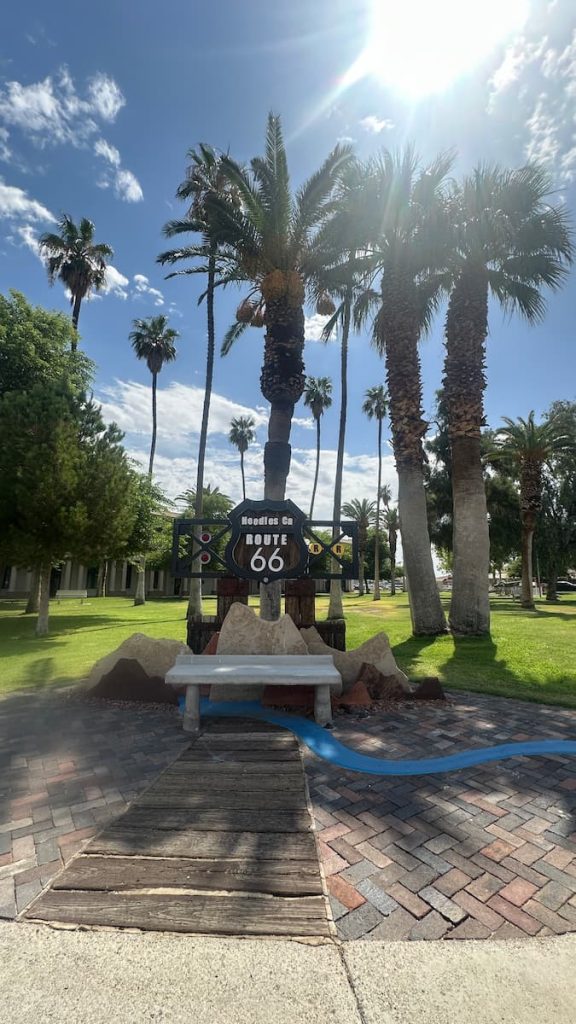 Un banc en pierre blanche, surmontée d'un panneau Route 66, devant des palmiers à Needles sur la route 66
