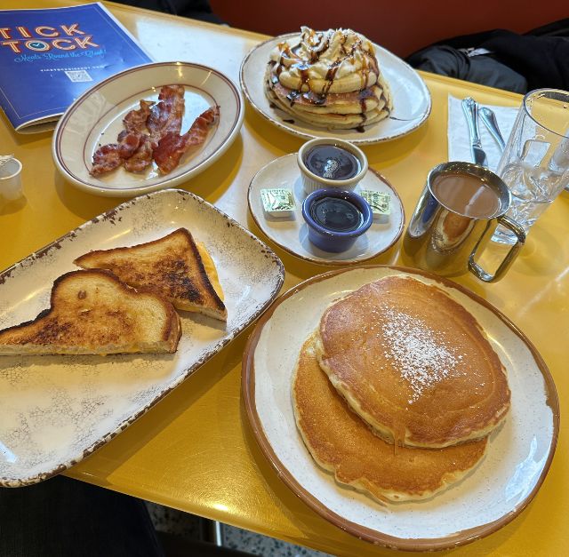Photo d'une table jaune avec plusieurs assiettes contenant des pancakes, des toasts, du bacon, une tasse de café et un chocolat chaud, à côté de la carte de restaurant "Tick Tock Diner" à New York
