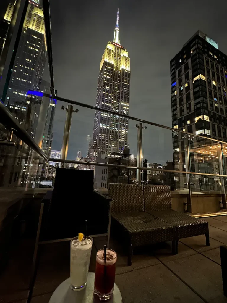 Deux verres d'apéritif, posé sur une table, sur un roof top avec vue sur l'Empire State Building illuminé en blanc, à New York