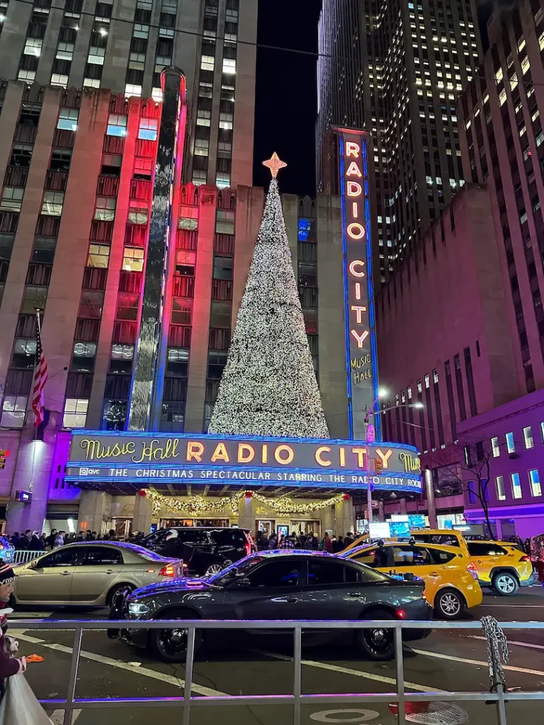 Photo prise de l'autre côté de la rue du radio city music hall, avec son sapin de Noël illuminé en vert, et un taxi jaune passant devant, à New York