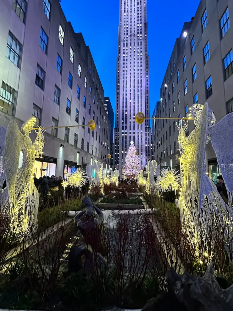 Photo de l'allée du Rockefeller Center Channel Gardens, avec le sapin de Noël illuminé en fond, et les angelots jouant de la trompette doré au premier plan, à New York