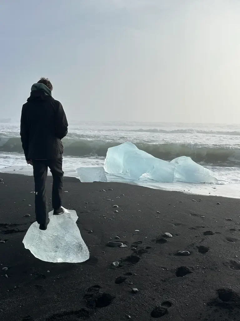 adolescent debout de dos sur un morceau d'iceberg scintillant sur le sable noir de la plage de diamond Beach, près de la lagune glacière de Jökulsárlón dans le sud de l'Islande