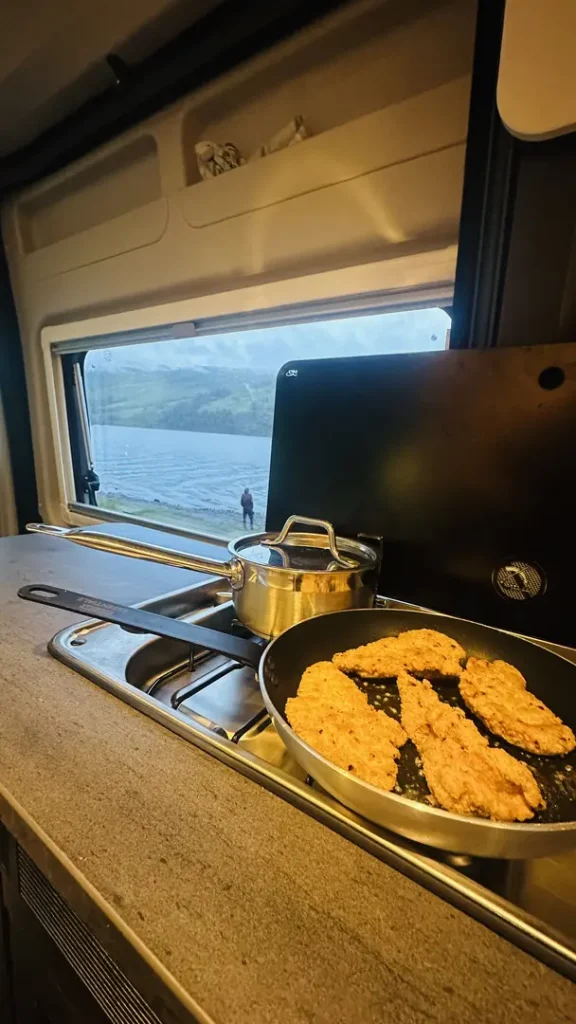 Une poëlle sur le feu avec du pulet devant la fenêtre avec vue sur le Loch Earn dans un van aménagé, en Ecosse, Royaume Uni